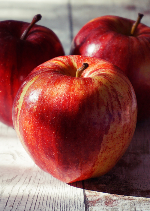 Three shiny red apples with subtle yellow streaks are placed on a rustic wooden surface, capturing sunlight and creating a fresh, inviting atmosphere.