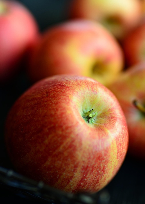 Close-up of several red apples with a glossy texture, centered on one apple with subtle yellow streaks. The background is softly blurred, creating a fresh, appetizing feel.