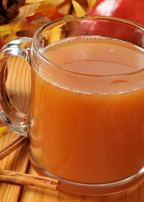 A clear glass mug filled with warm apple cider sits on a wooden table, surrounded by cinnamon sticks and autumn leaves, evoking a cozy, fall ambiance.