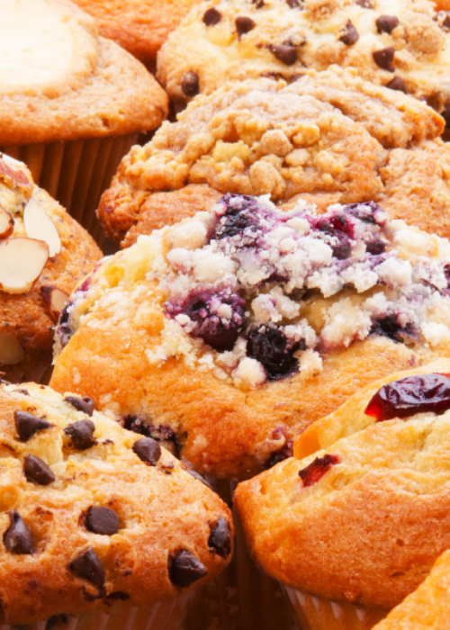 A close-up of assorted muffins with toppings like blueberries, chocolate chips, almonds, and crumble. The image conveys a warm, inviting tone.