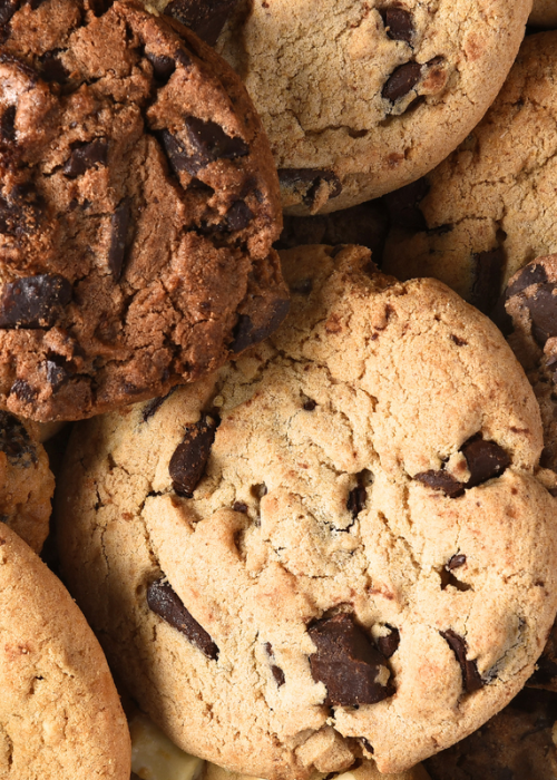 A close-up of assorted chocolate chip cookies, showcasing their golden-brown texture and large chocolate chunks. The image evokes a warm, inviting feel.