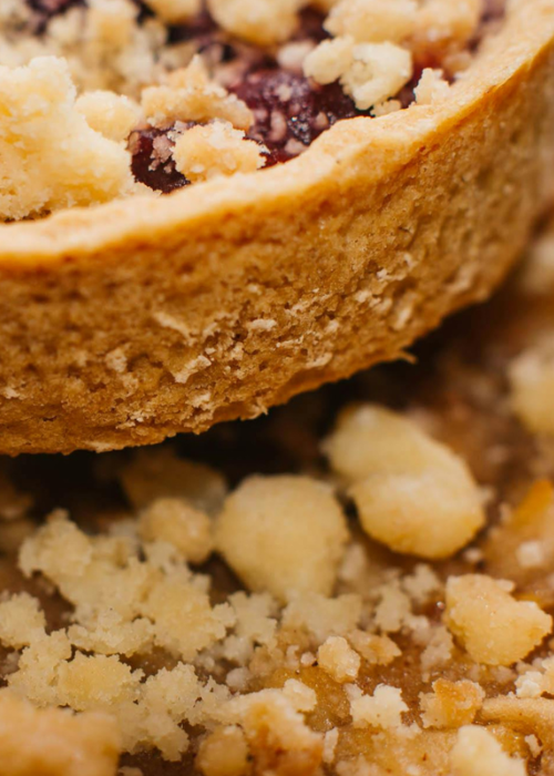 Close-up of a crumbled pastry, showcasing a textured, golden crust with a rich berry filling. Crumbs scatter around, conveying a freshly baked feel.
