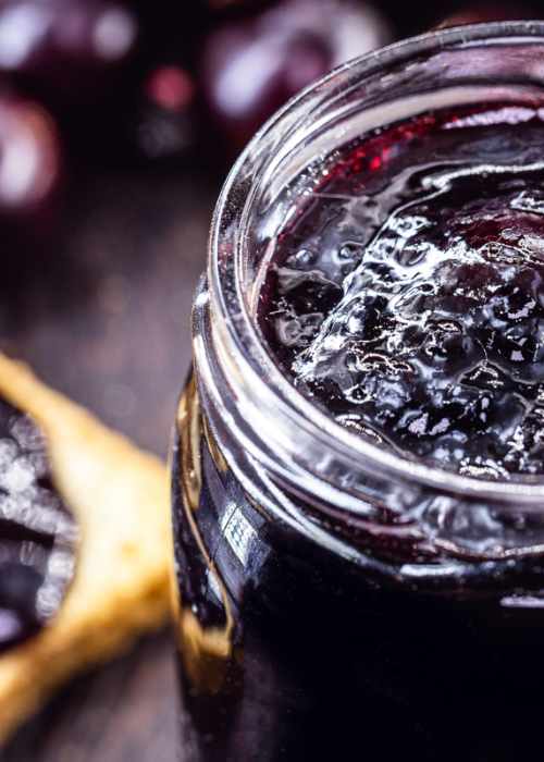 Close-up of a jar filled with deep red cherry preserves on a dark wooden surface, with blurred cherries and a crispy cracker as background accents.