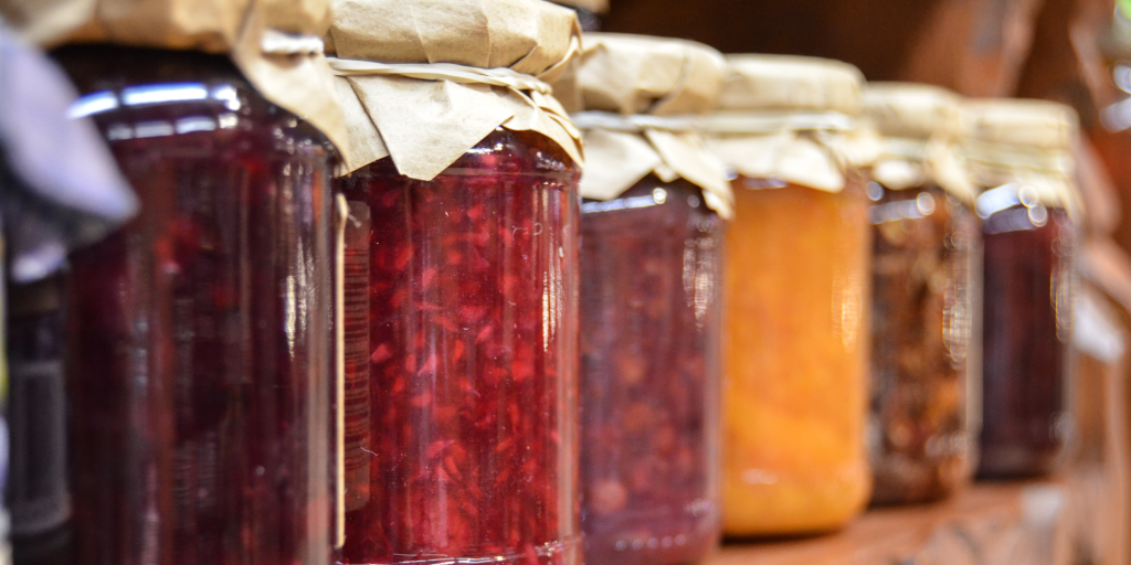 A row of colorful jam jars on a wooden shelf, featuring red, orange, and brown hues. They are sealed with paper covers, evoking a rustic, homemade feel.